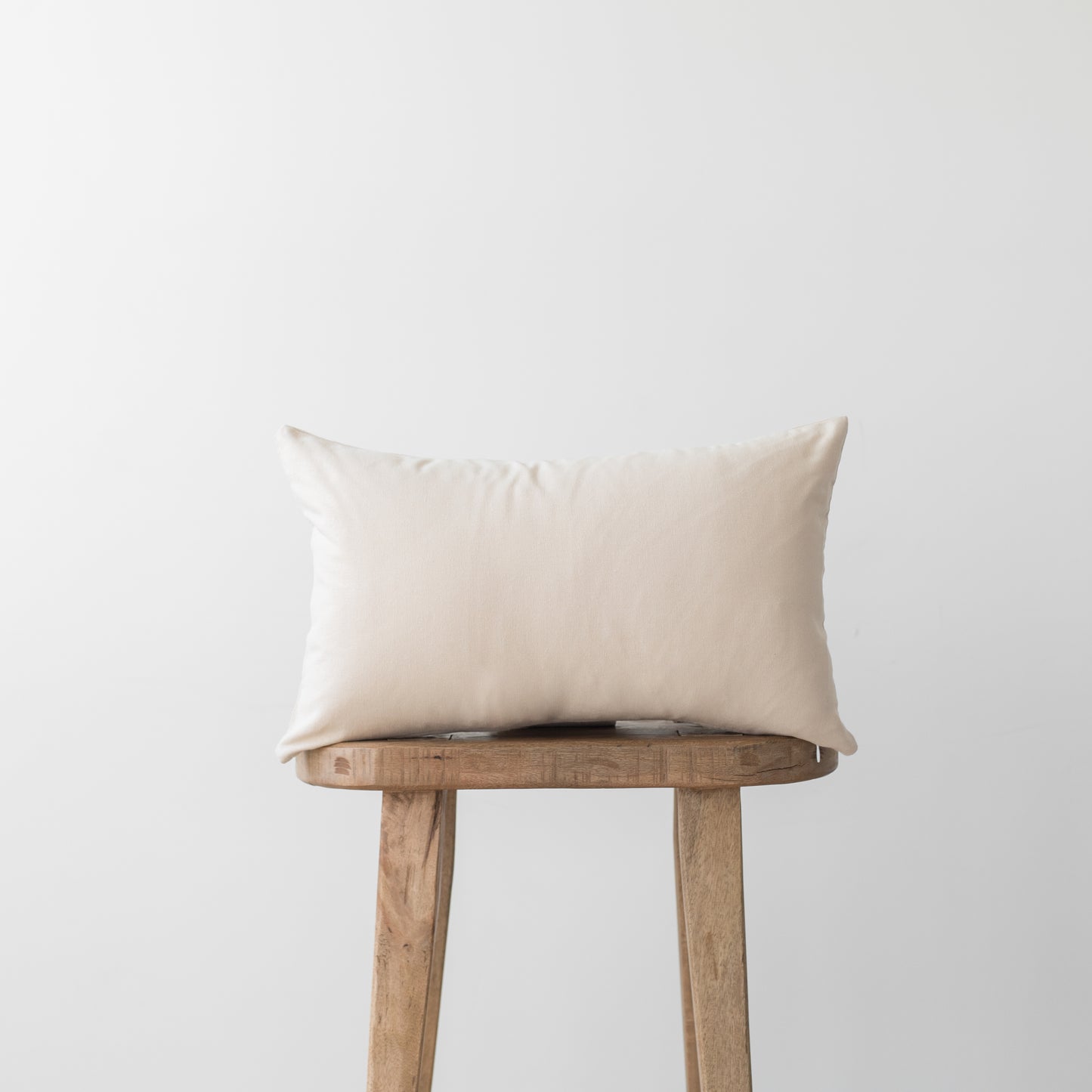 Cream-colored rectangular pillow with smooth fabric displayed on a wooden stool.