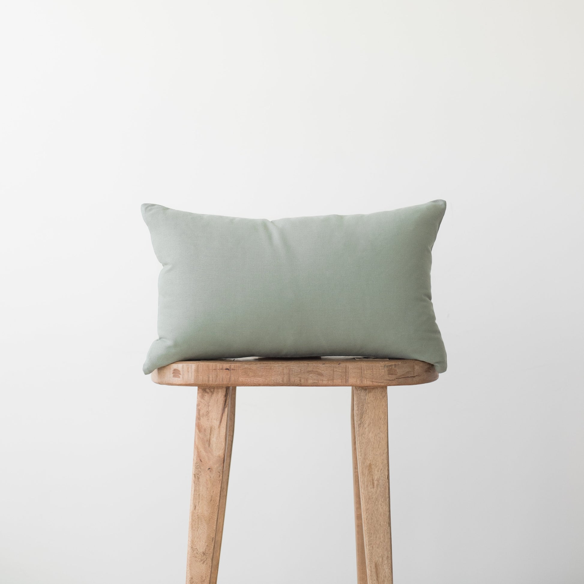 Sage green rectangular pillow with solid woven fabric displayed on wooden stool.