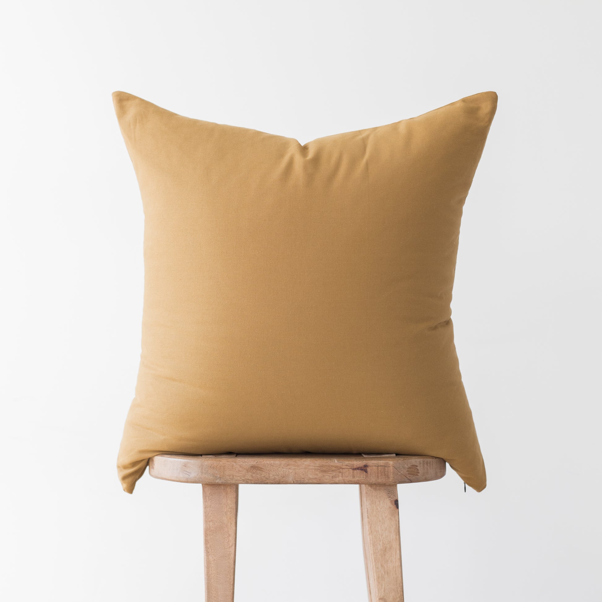 Gold cushion cover displayed on wooden stool against light gray background.