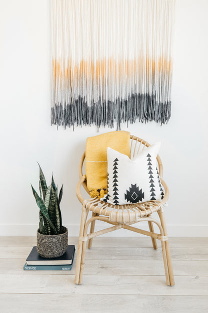 Woven wall hanging with cream, mustard, and charcoal ombre stripes displayed above a rattan chair.