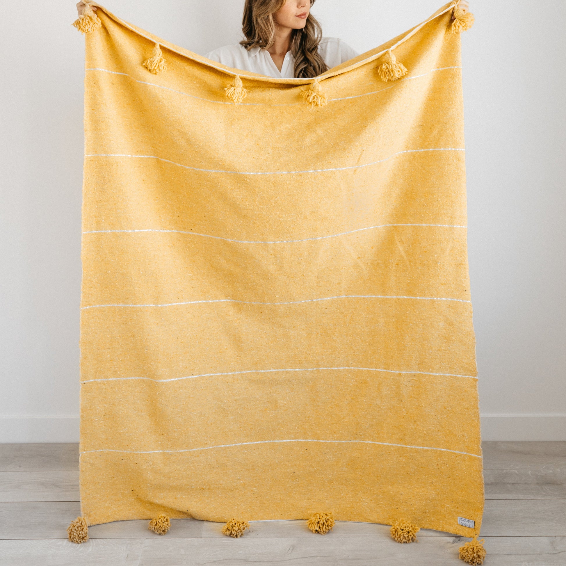Gold throw blanket with horizontal stripes, tassels at corners, held by woman in white shirt.