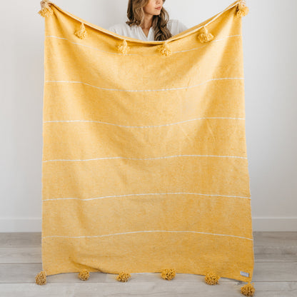 Gold throw blanket with horizontal stripes, tassels at corners, held by woman in white shirt.