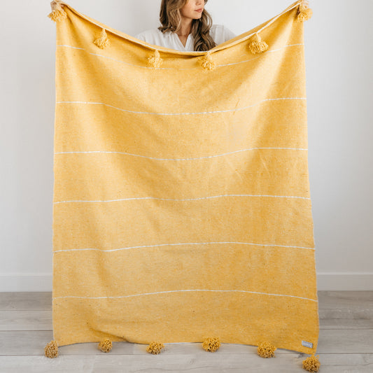 Gold throw blanket with horizontal stripes, tassels at corners, held by woman in white shirt.