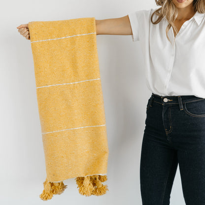 Yellow woven scarf with white stripes and tassel fringe ends, held by woman wearing white shirt and dark jeans.