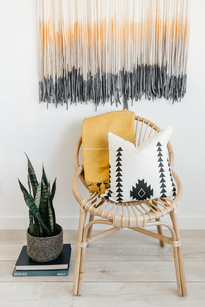 Woven rattan chair with yellow throw blanket and geometric patterned pillow, styled with potted plant and wall hanging.