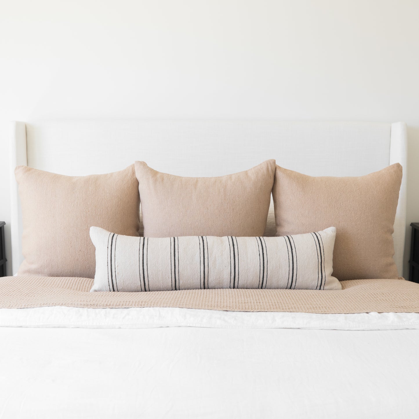 Bed styled with three beige linen pillows and one striped lumbar pillow against white headboard.