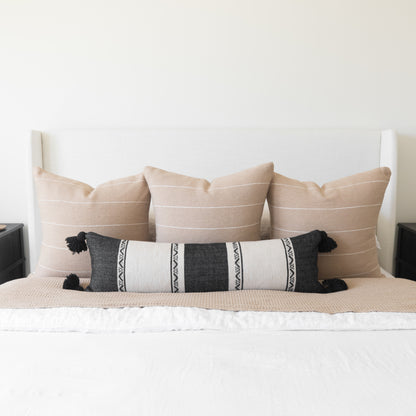 Charcoal and cream striped lumbar pillow with black tassels displayed on a bed with tan pillows.