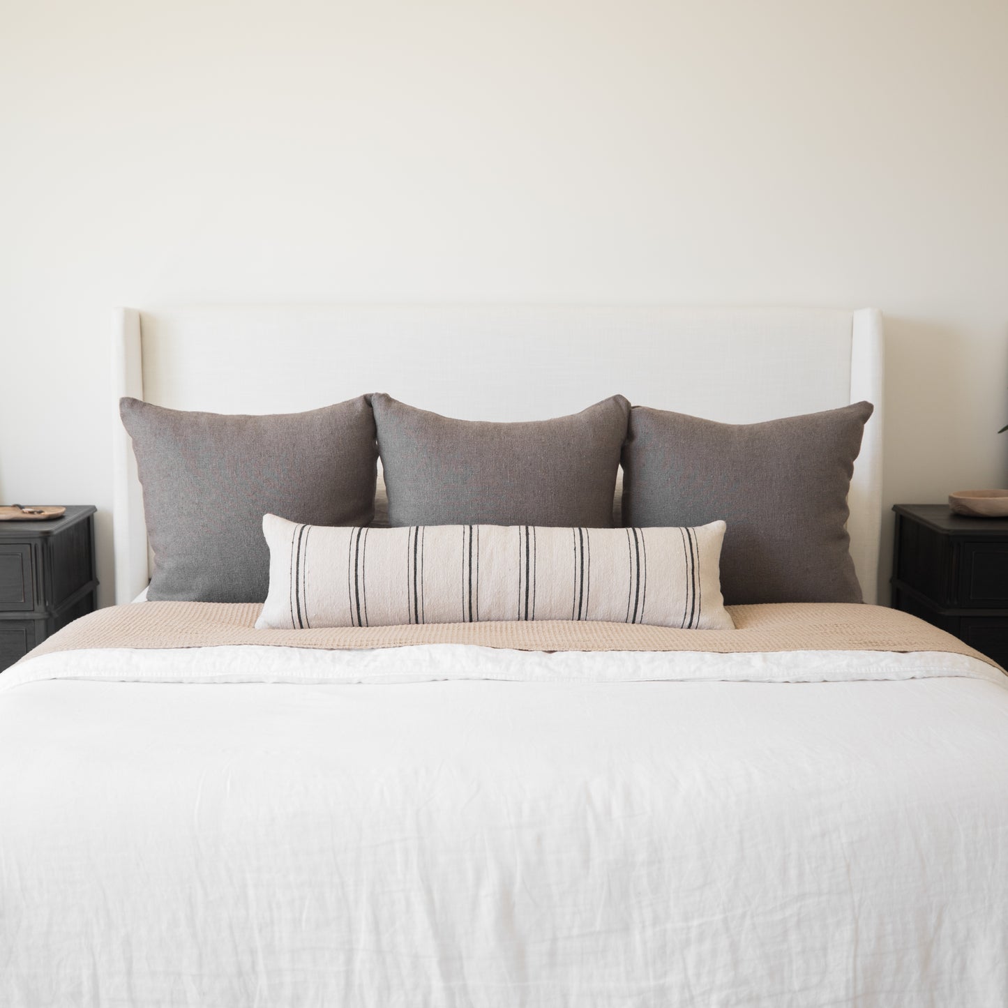 Bed with white upholstered headboard, white bedding, tan quilt, three gray pillows, and one striped lumbar pillow.