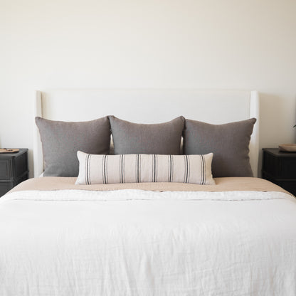 Bed with white upholstered headboard, white bedding, tan quilt, three gray pillows, and one striped lumbar pillow.