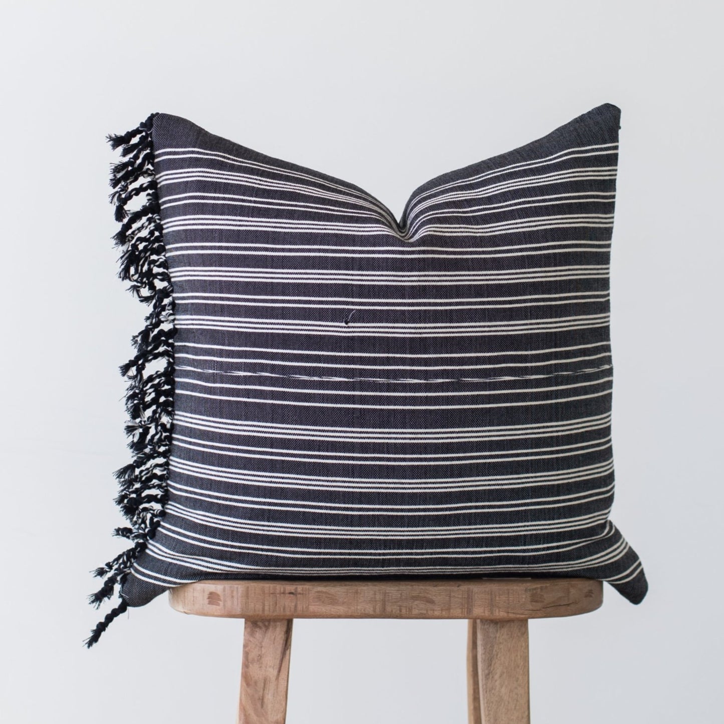 Dark gray pillow with horizontal white stripes and black fringe edges, displayed on wooden stool.