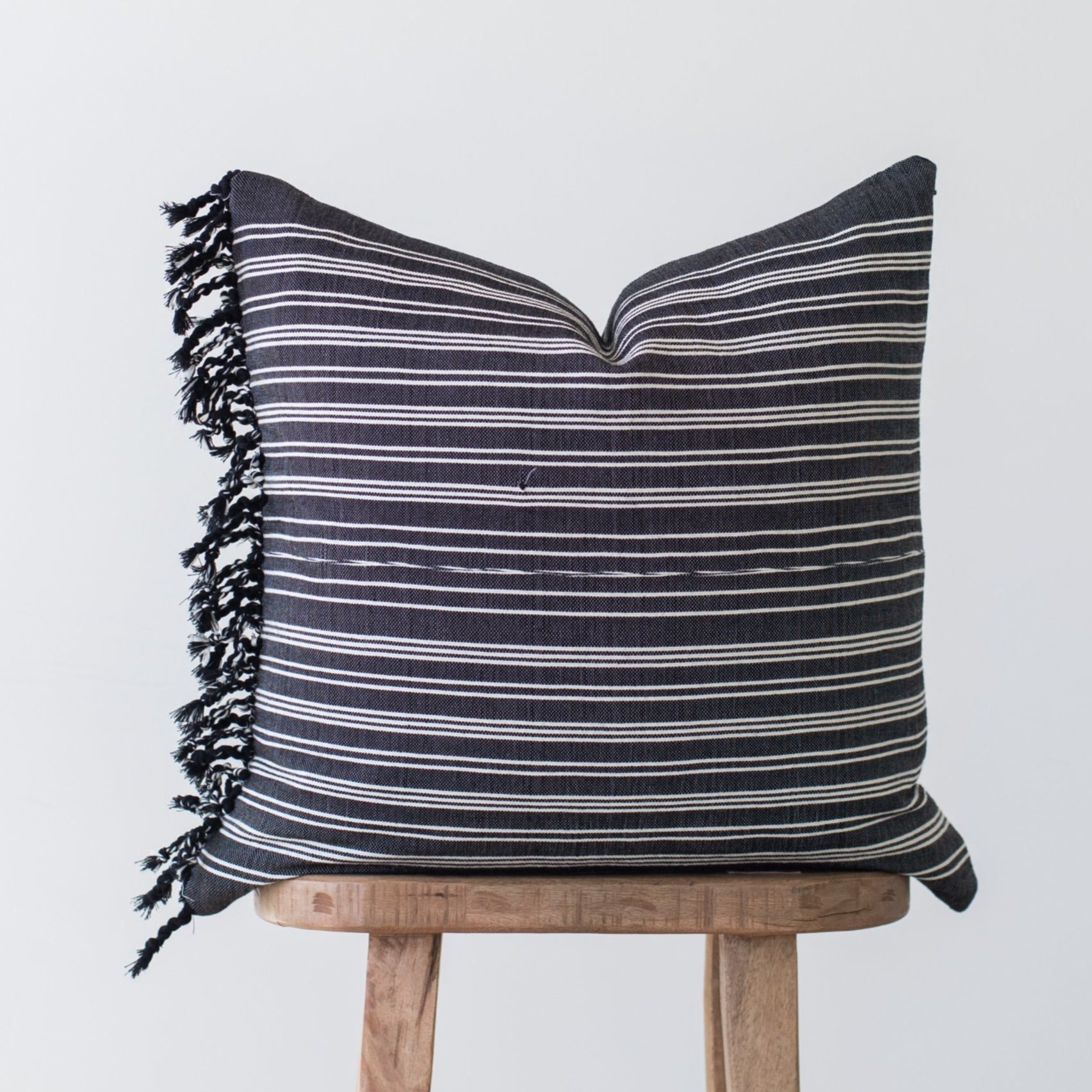 Dark gray pillow with horizontal white stripes and black fringe edges, displayed on wooden stool.