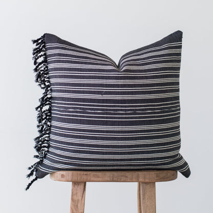 Dark gray pillow with horizontal white stripes and black fringe edges, displayed on wooden stool.