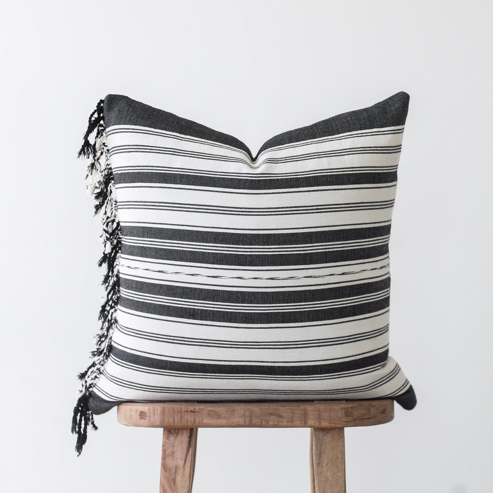 Black and white striped pillow with tasseled edges on wooden stool.