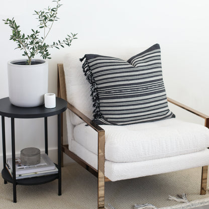 Black and white striped pillow with fringe trim on natural wood-frame chair with cream cushion.