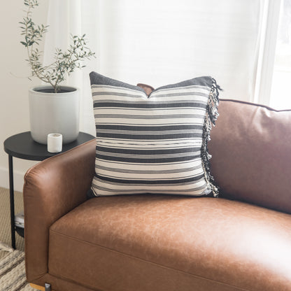 Striped throw pillow with fringe trim in charcoal and cream on a brown leather chair.