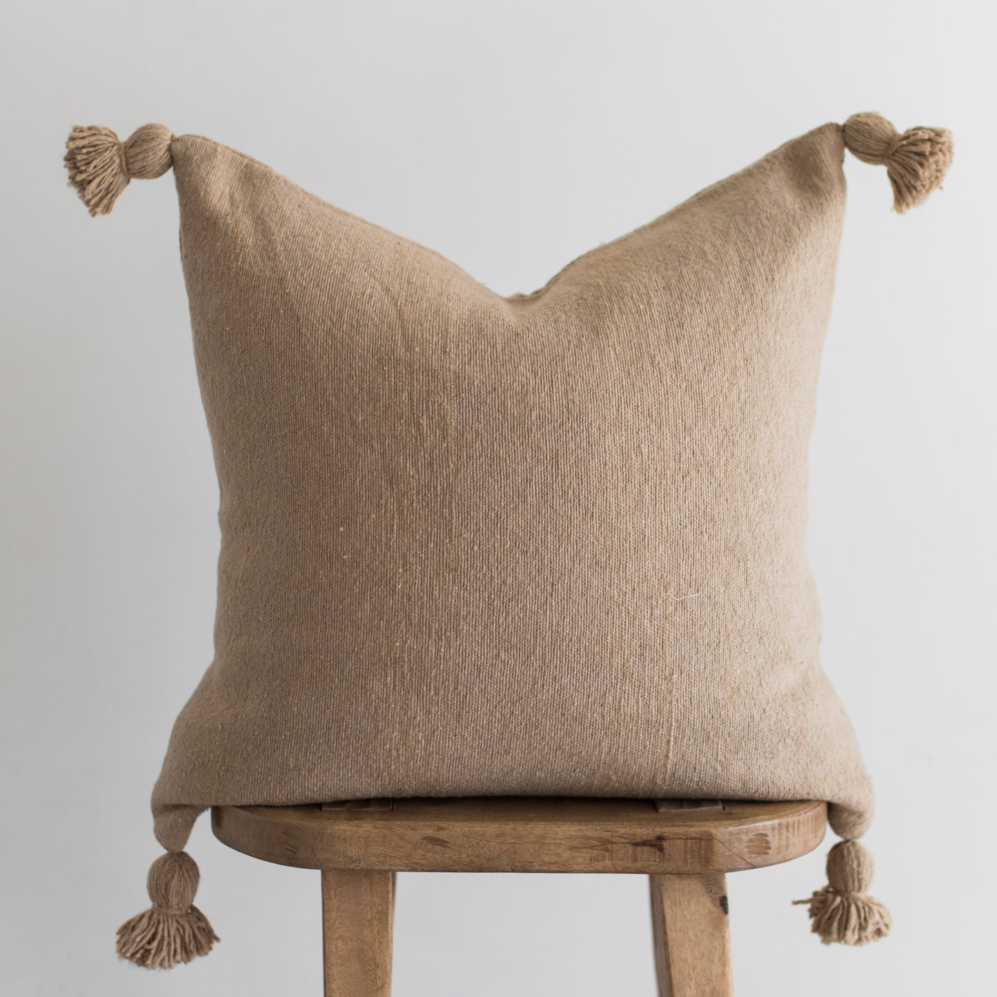 Beige linen throw pillow with knotted tassels on corners, displayed on wooden stool.