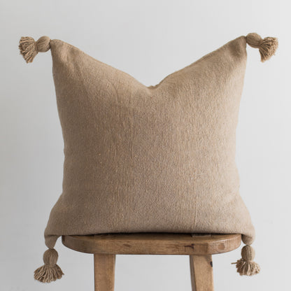 Beige linen throw pillow with knotted tassels on corners, displayed on wooden stool.