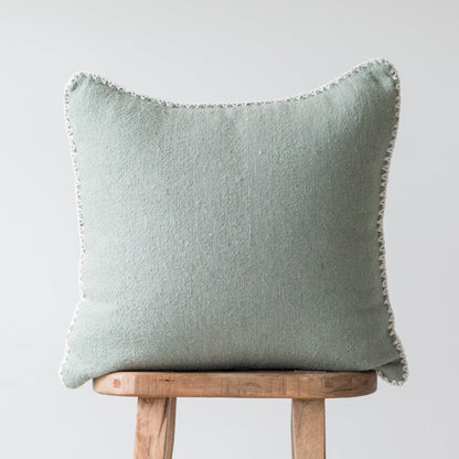 Sage green square pillow with frayed edges resting on wooden chair back.
