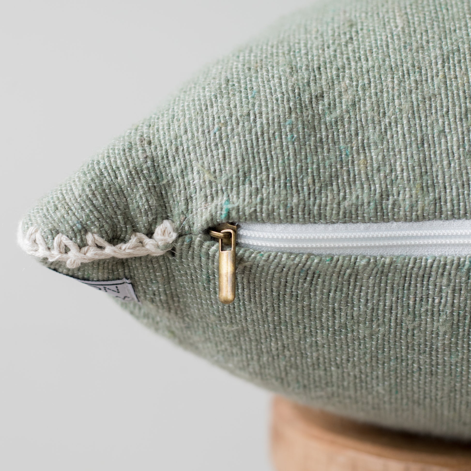 Sage green 22-inch pillow with white zipper, cream rope trim, and gold slider detail on textured fabric.