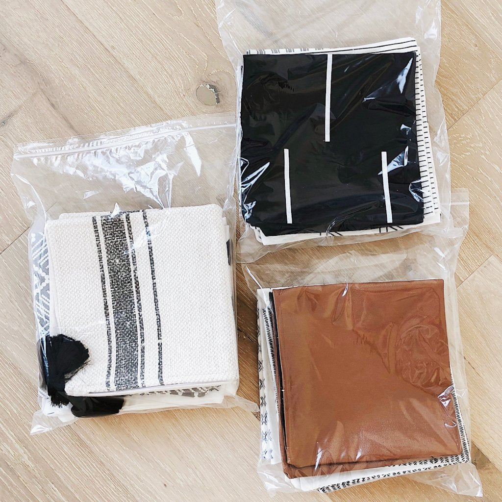 Six-pack grab bag containing white striped cloth, black shorts, brown leather item, and clear plastic pouches on wooden surfa