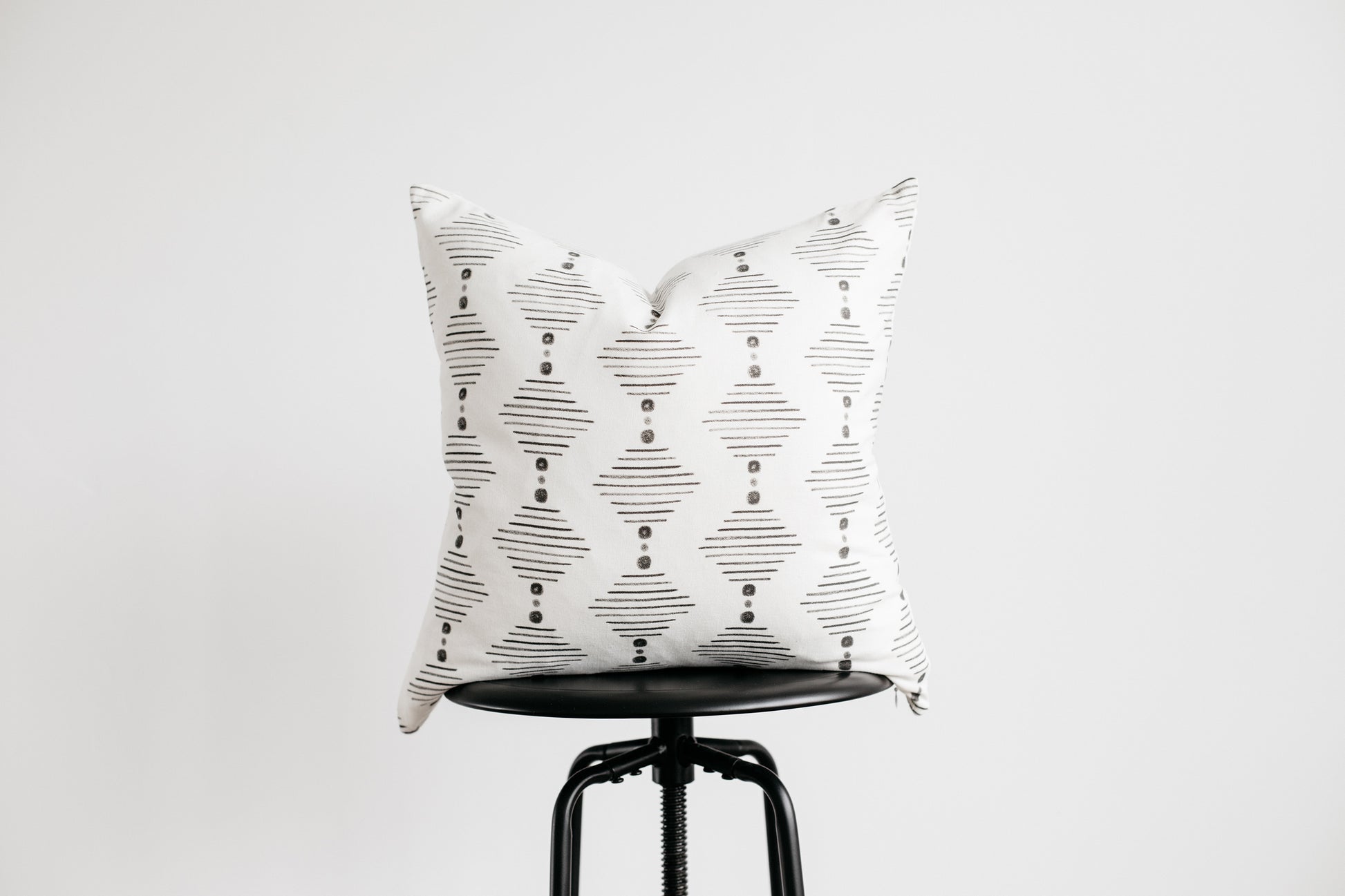White square pillow with black horizontal line pattern and dots, displayed on black stool.