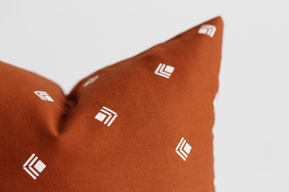 Rust-colored pillow with white geometric logo pattern scattered across fabric surface.
