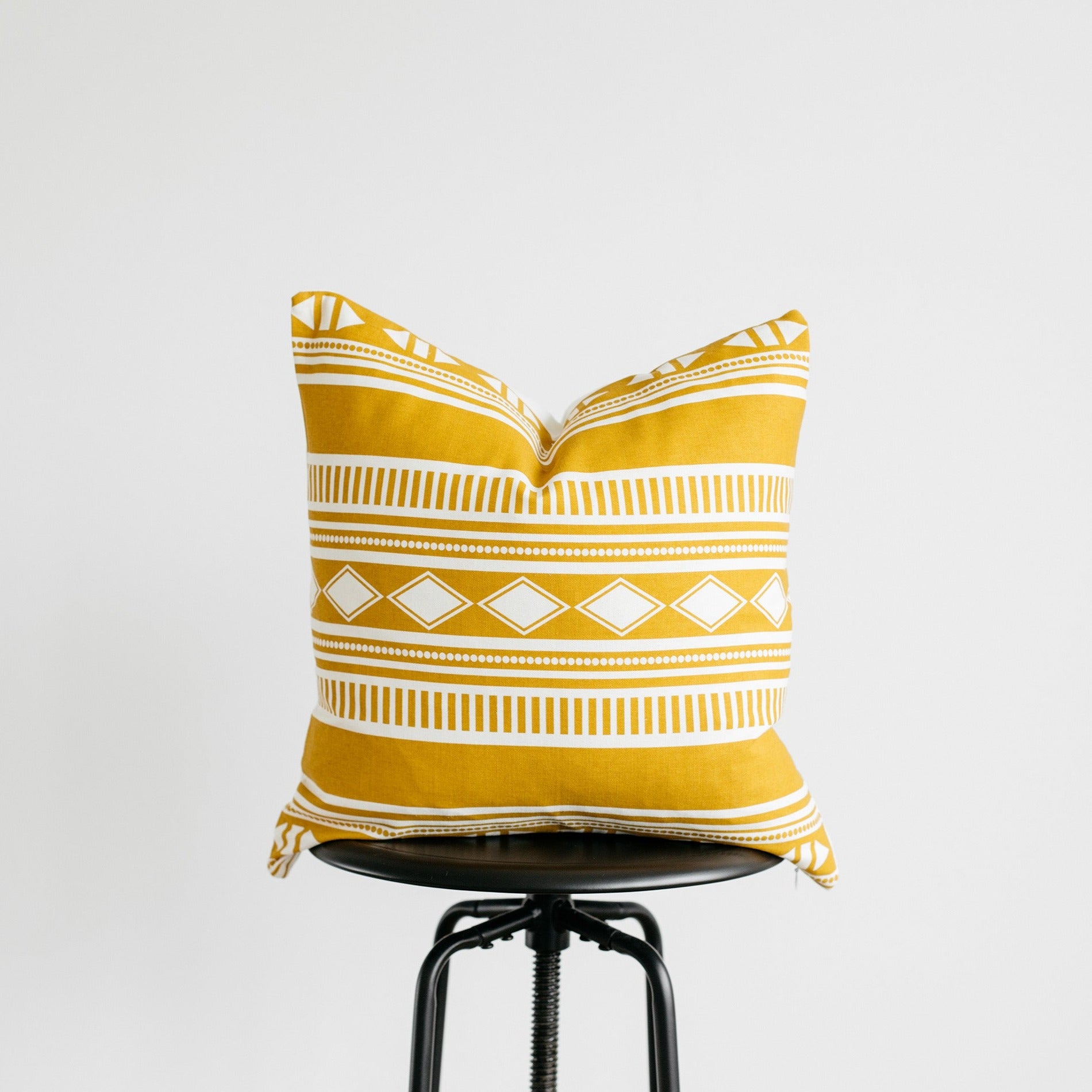 Gold and white geometric patterned cushion cover with striped and diamond motifs displayed on a black stool.