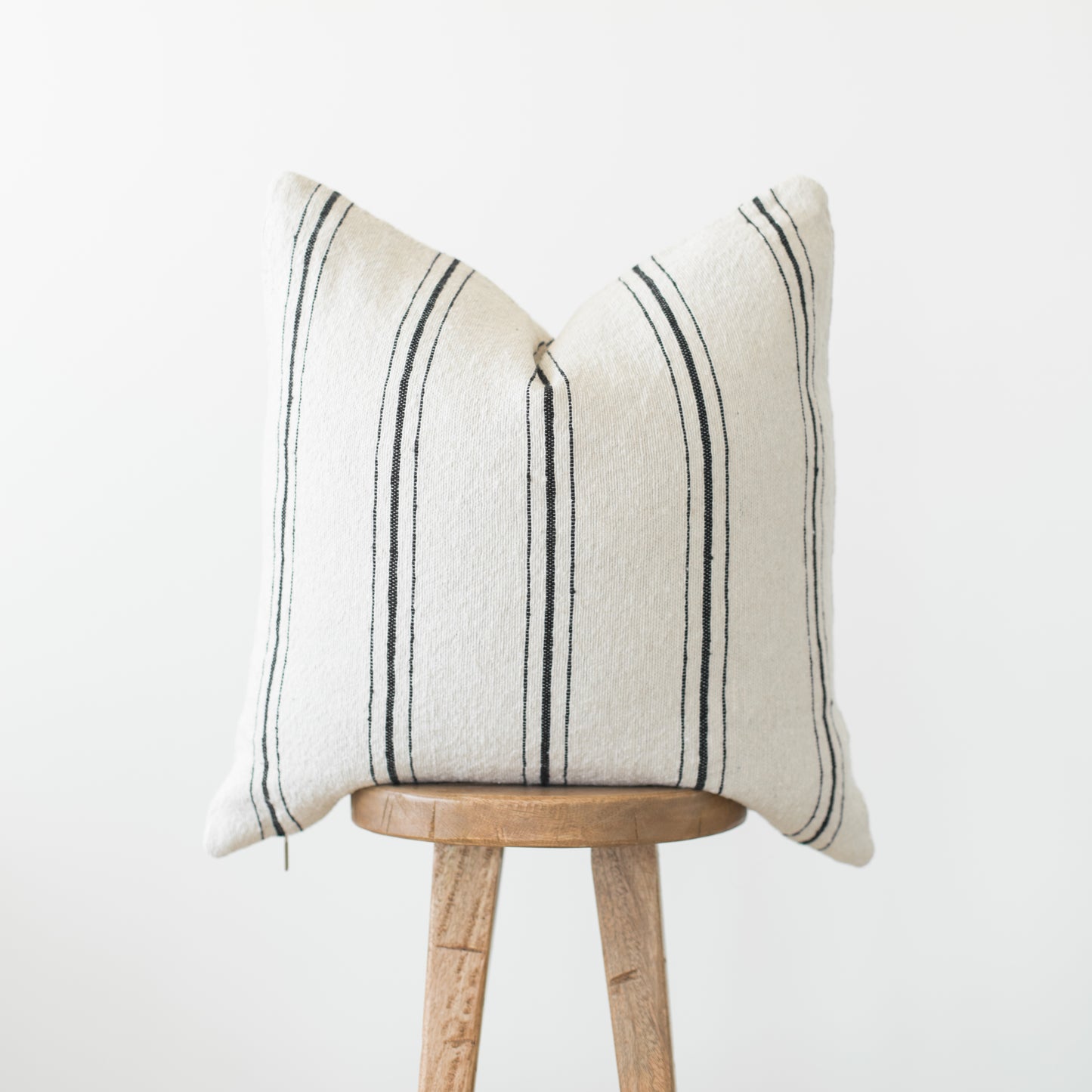 Cream pillow with dark vertical stripes displayed on wooden stool against white background.