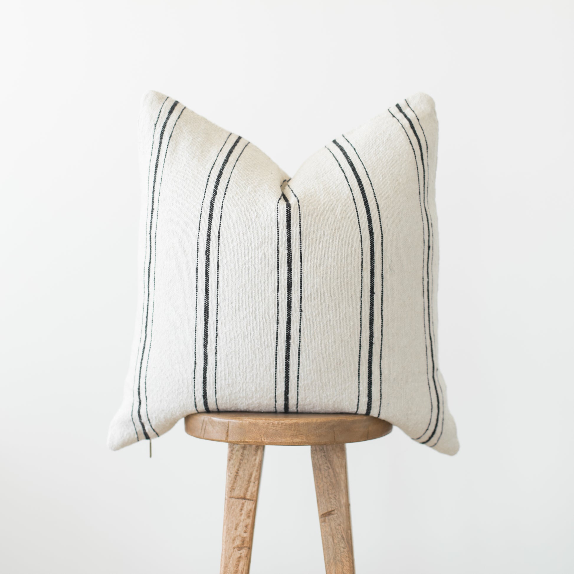 Cream pillow with dark vertical stripes displayed on wooden stool against white background.