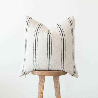 Cream pillow with dark vertical stripes displayed on wooden stool against white background.