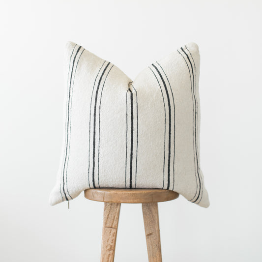 Cream pillow with dark vertical stripes displayed on wooden stool against white background.
