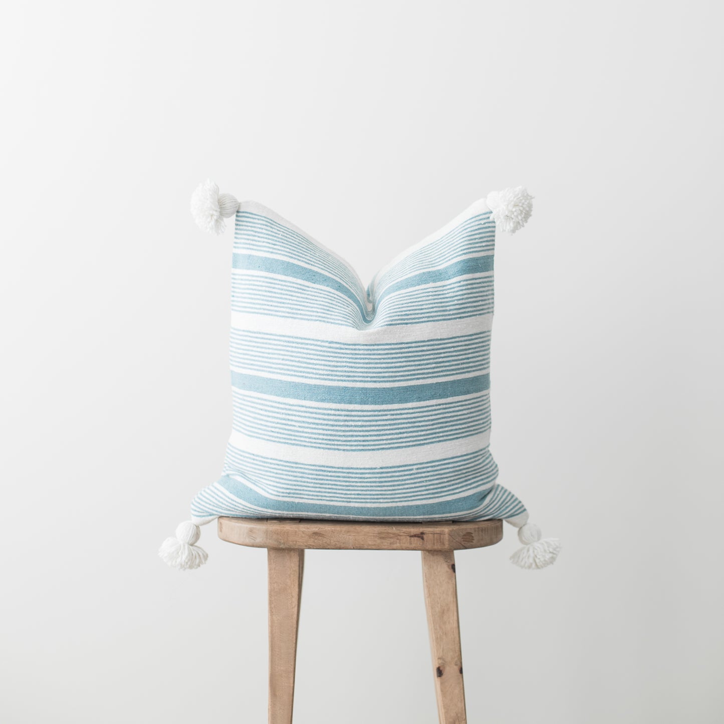 Blue and white striped throw pillow with white corner tassels on wooden stool.