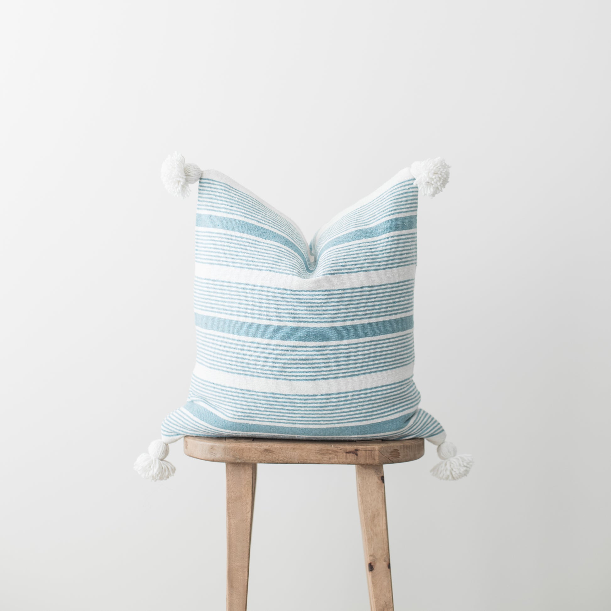 Blue and white striped throw pillow with white corner tassels on wooden stool.