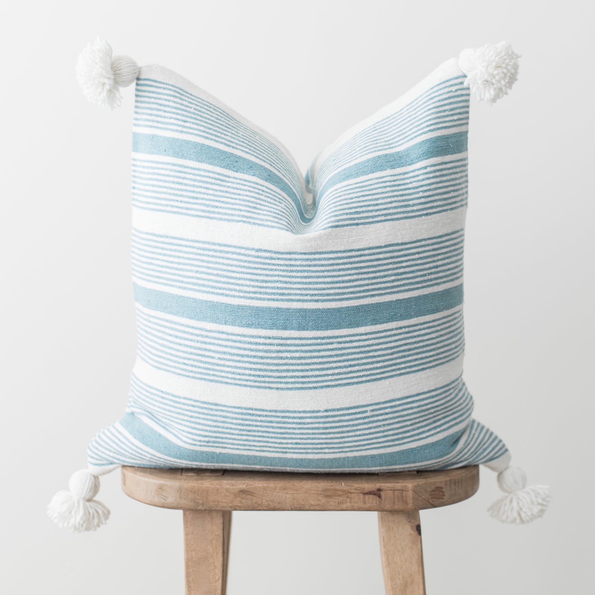 Blue and white striped pillow with white tassel corners displayed on wooden stool.