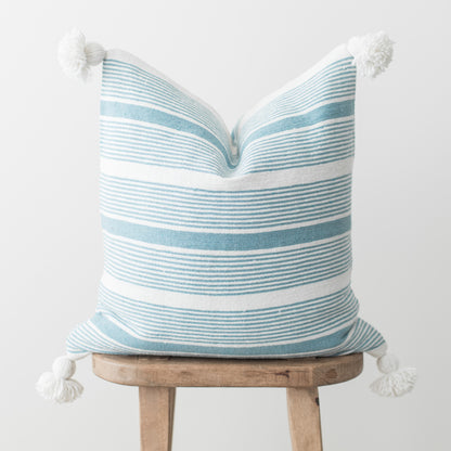 Blue and white striped pillow with white tassel corners displayed on wooden stool.