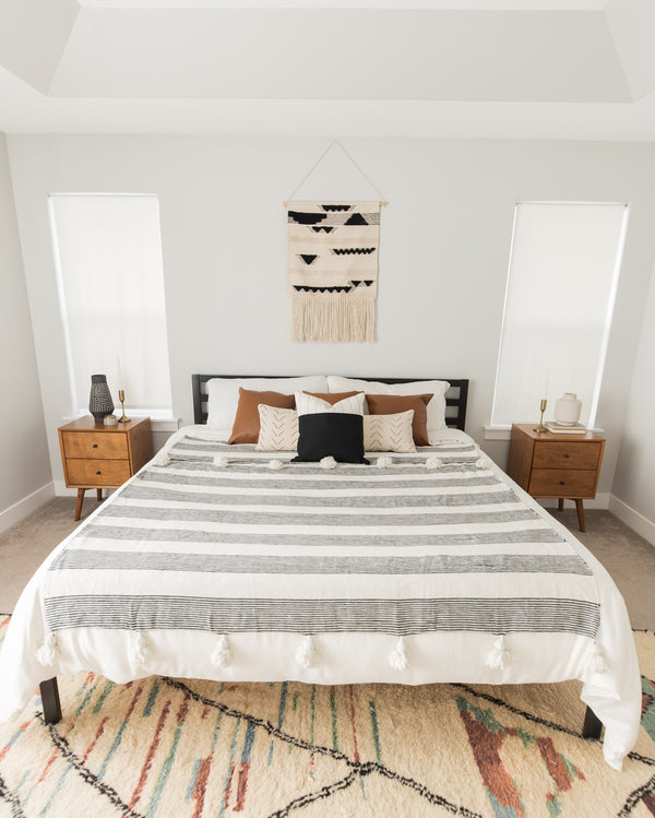 Large bed with gray and white striped bedding, tan and brown decorative pillows, and white pom-pom trim.