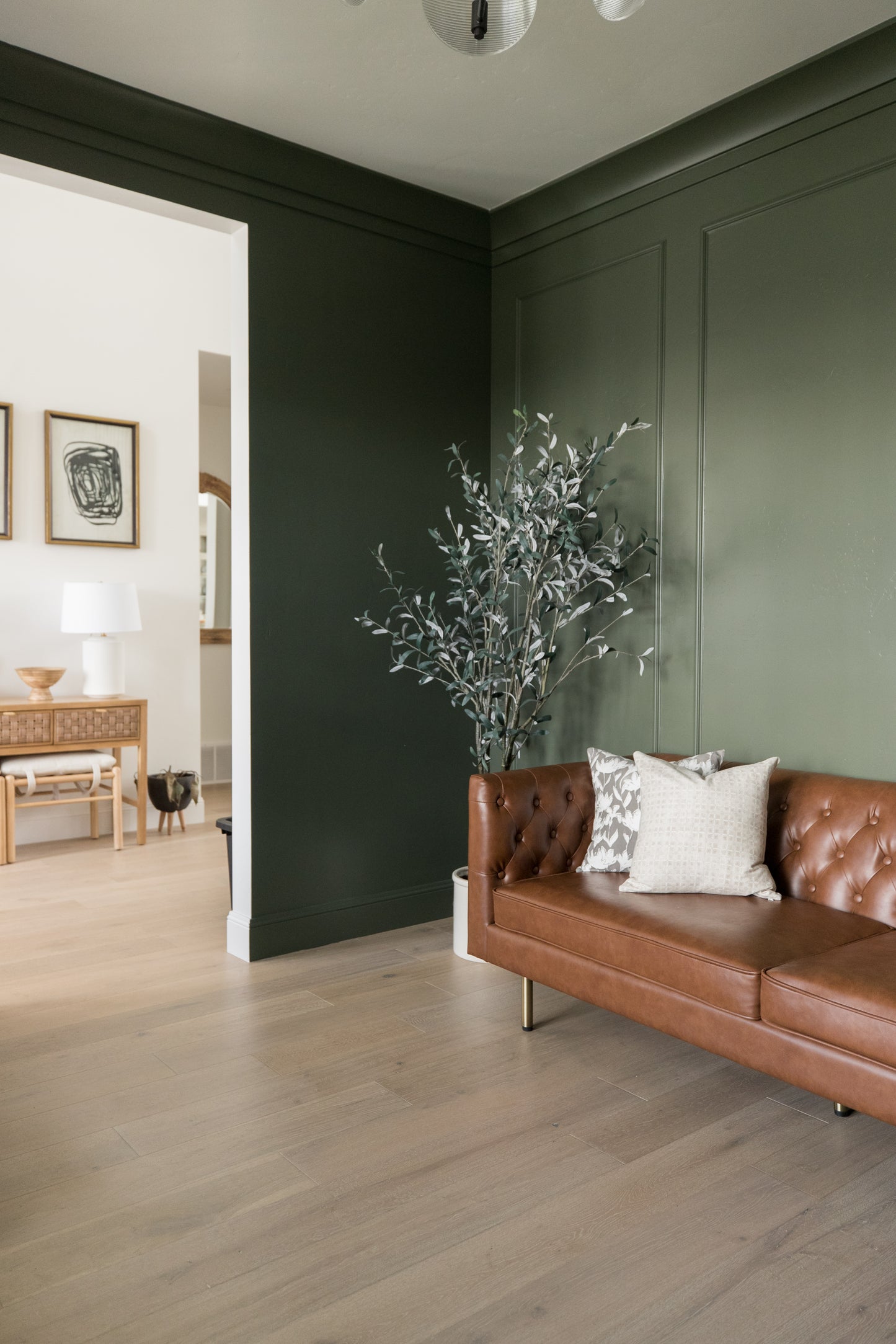 Dried greenery stems in terracotta vase displayed on brown leather sofa against dark green wall.