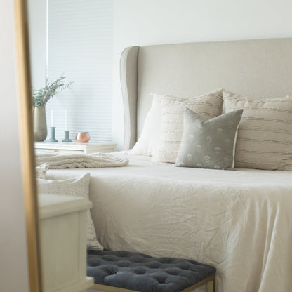 Gray floral throw pillow on a bed with cream bedding and upholstered headboard.