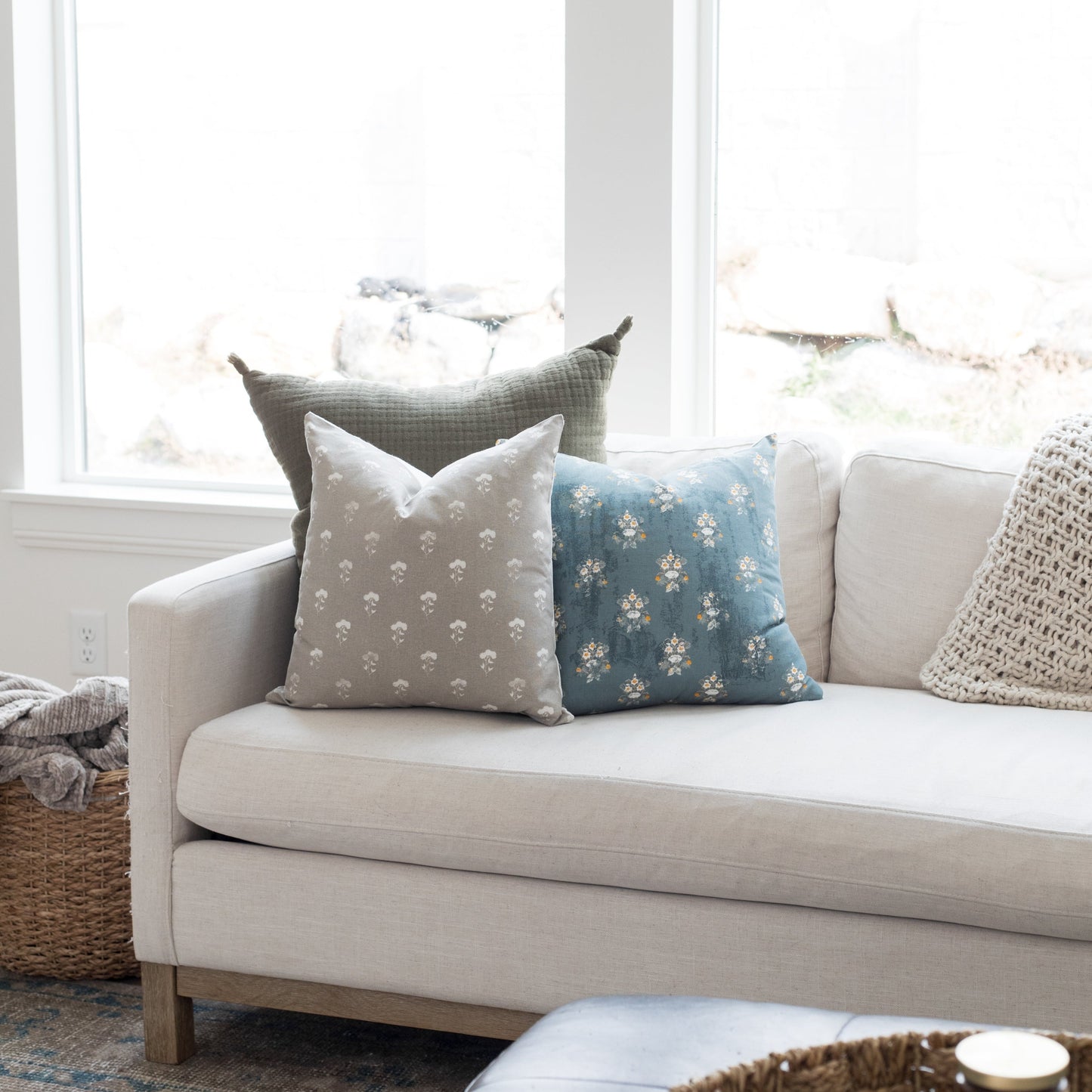 Gray and cream floral patterned pillow displayed on a beige sofa with other decorative pillows by a window.