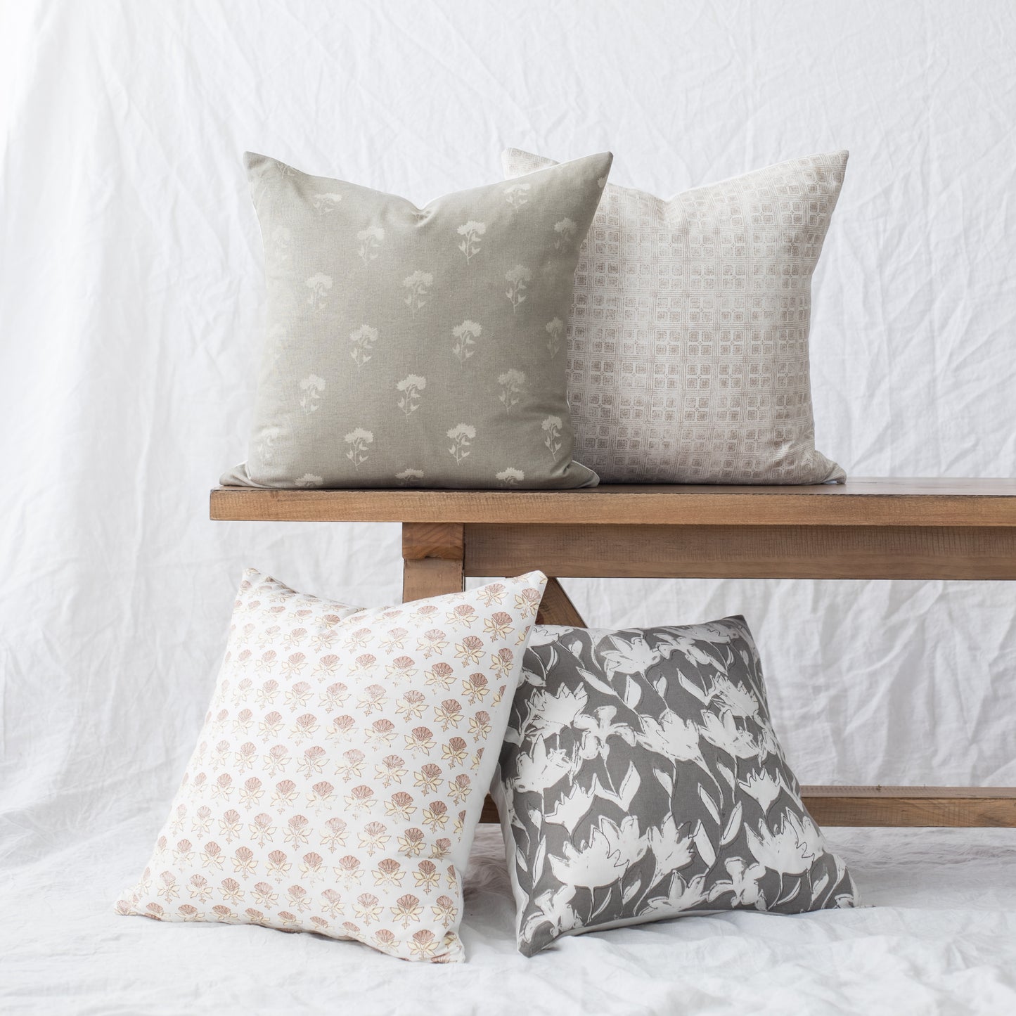 Four 18-inch square pillows with floral and geometric patterns in beige, taupe, and charcoal displayed on wooden shelves.