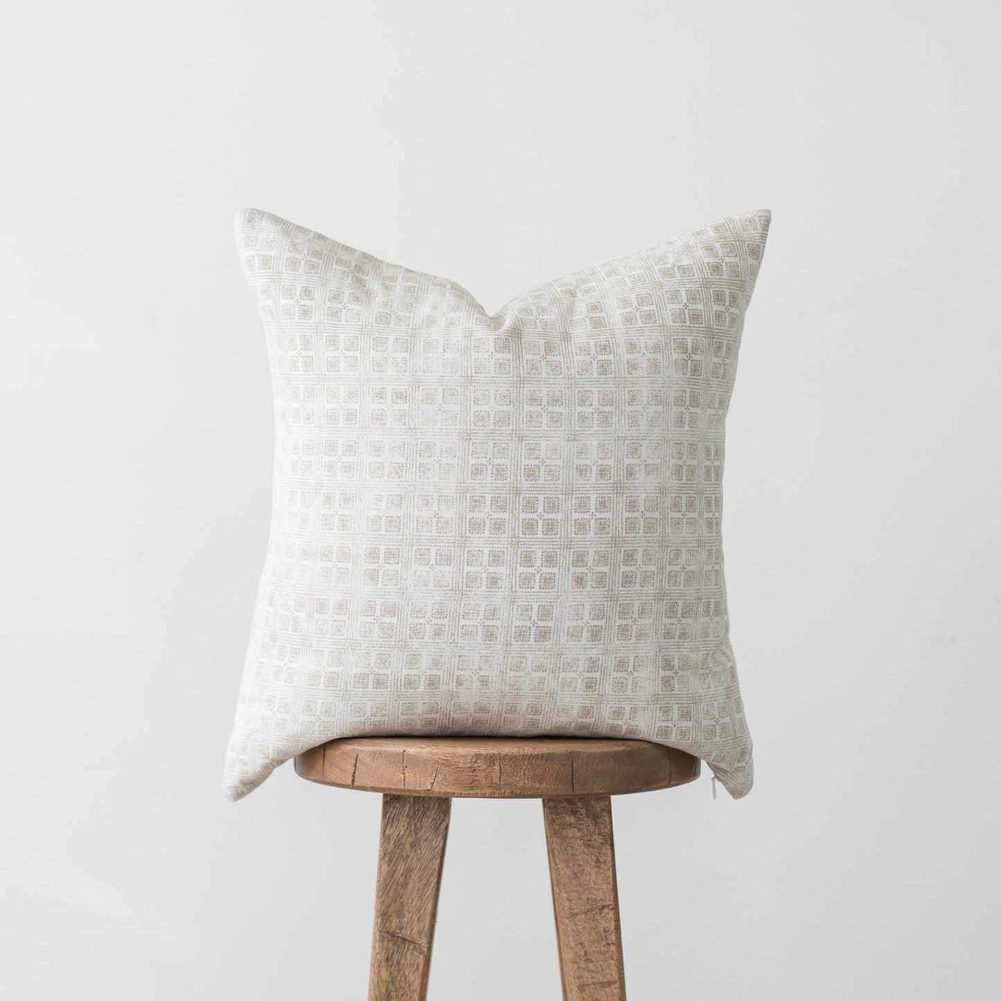 Cream-colored pillow with geometric grid pattern displayed on wooden stool against white background.