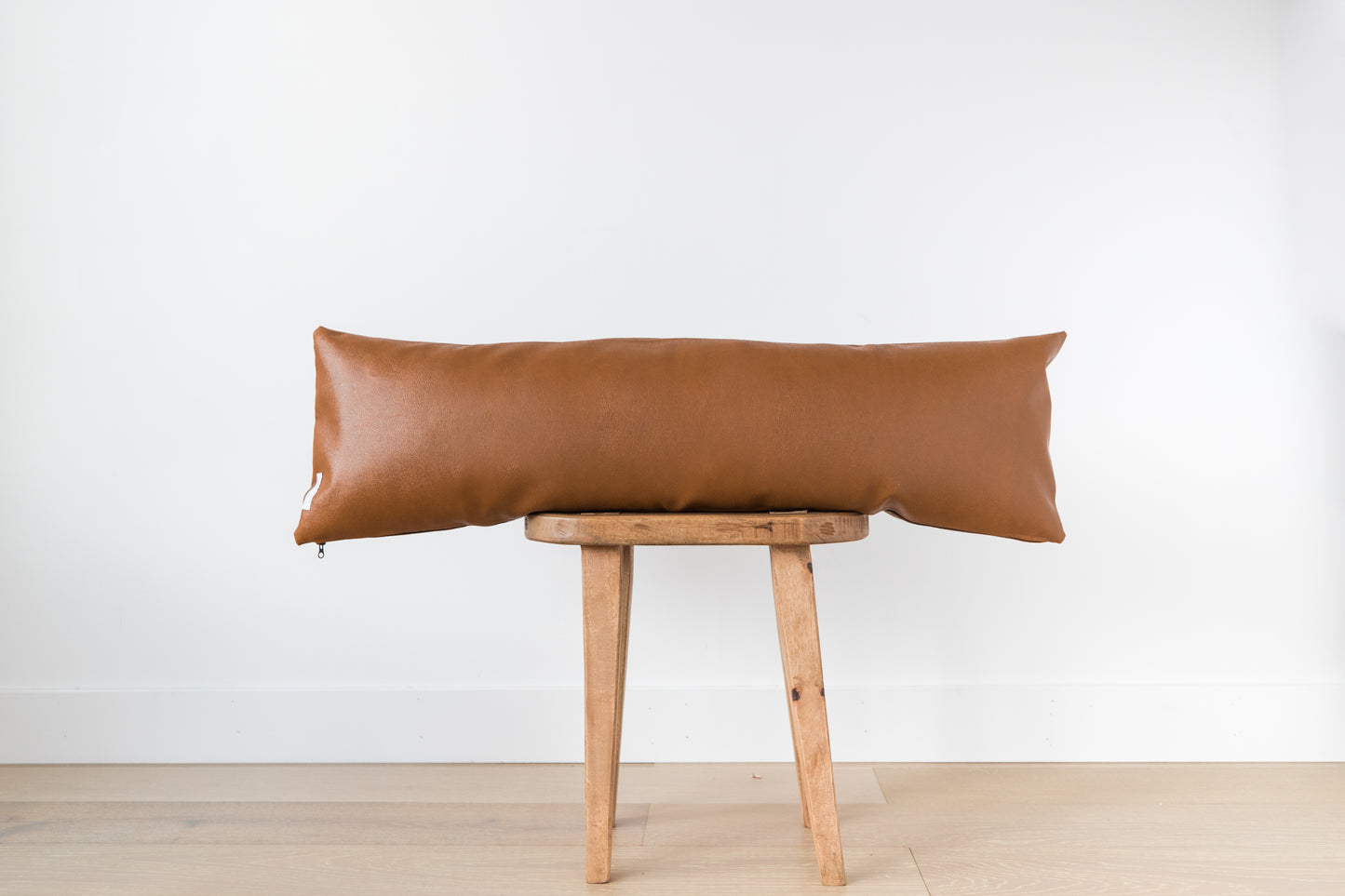 Brown leather lumbar pillow draped across wooden stool against white wall.