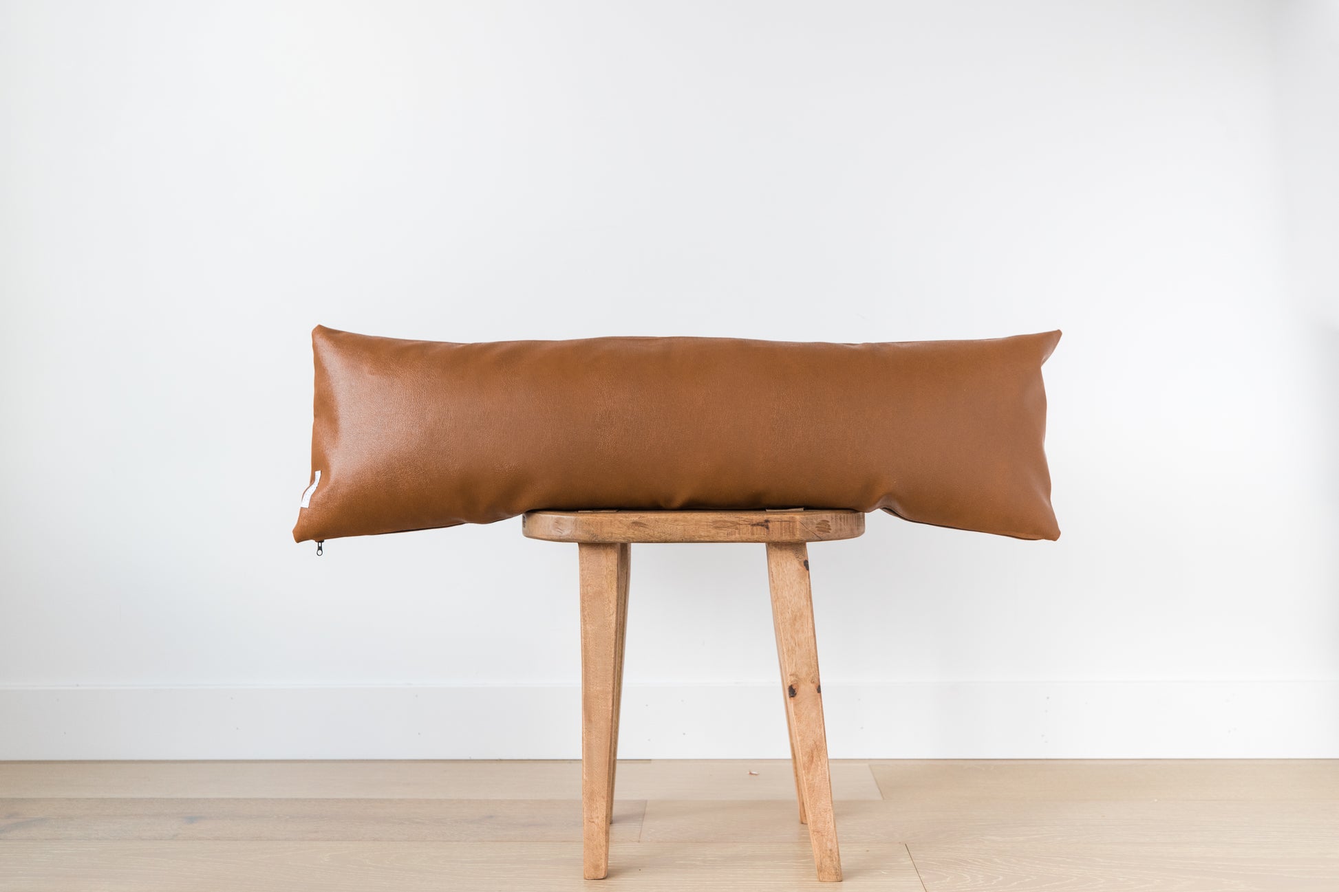 Brown leather lumbar pillow draped across wooden stool against white wall.