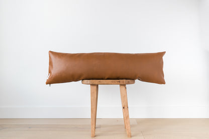 Brown leather lumbar pillow draped across wooden stool against white wall.