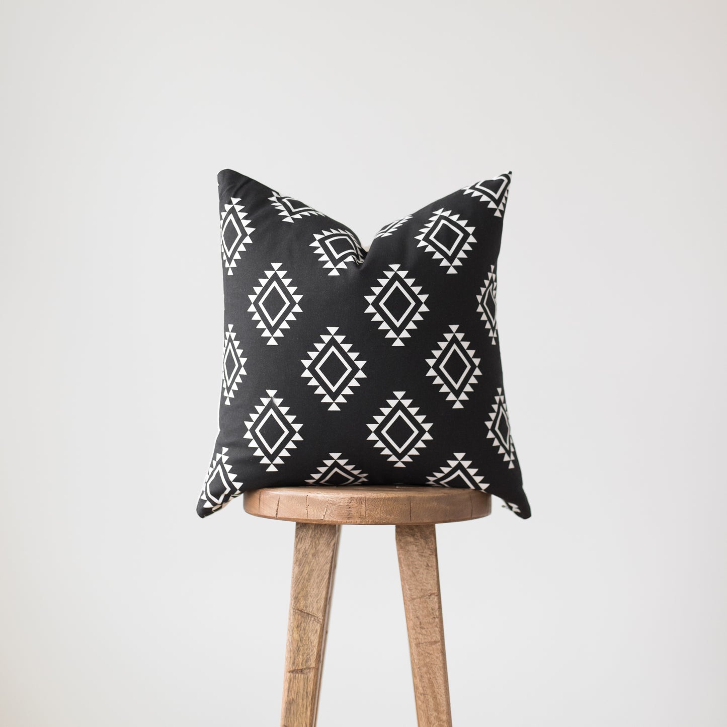 Black pillow with white geometric diamond pattern draped over wooden tripod stool.
