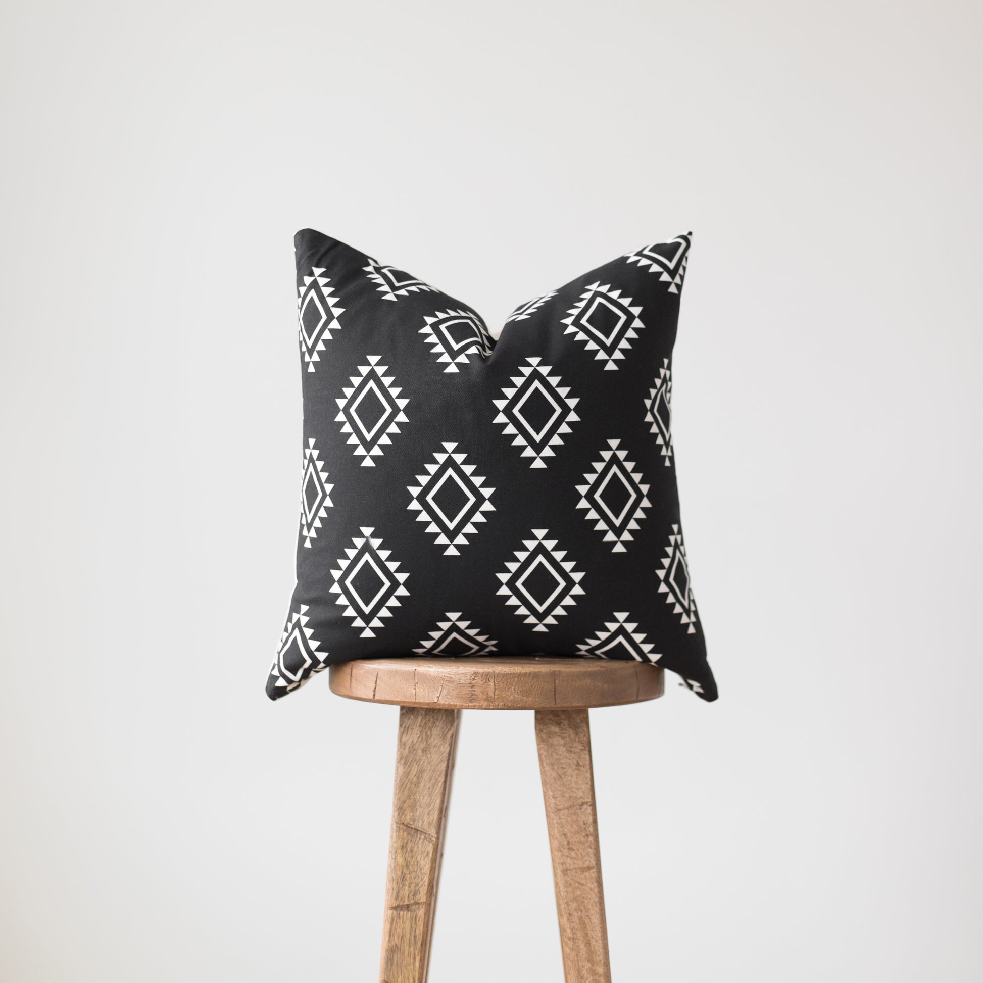 Black pillow with white geometric diamond pattern draped over wooden tripod stool.