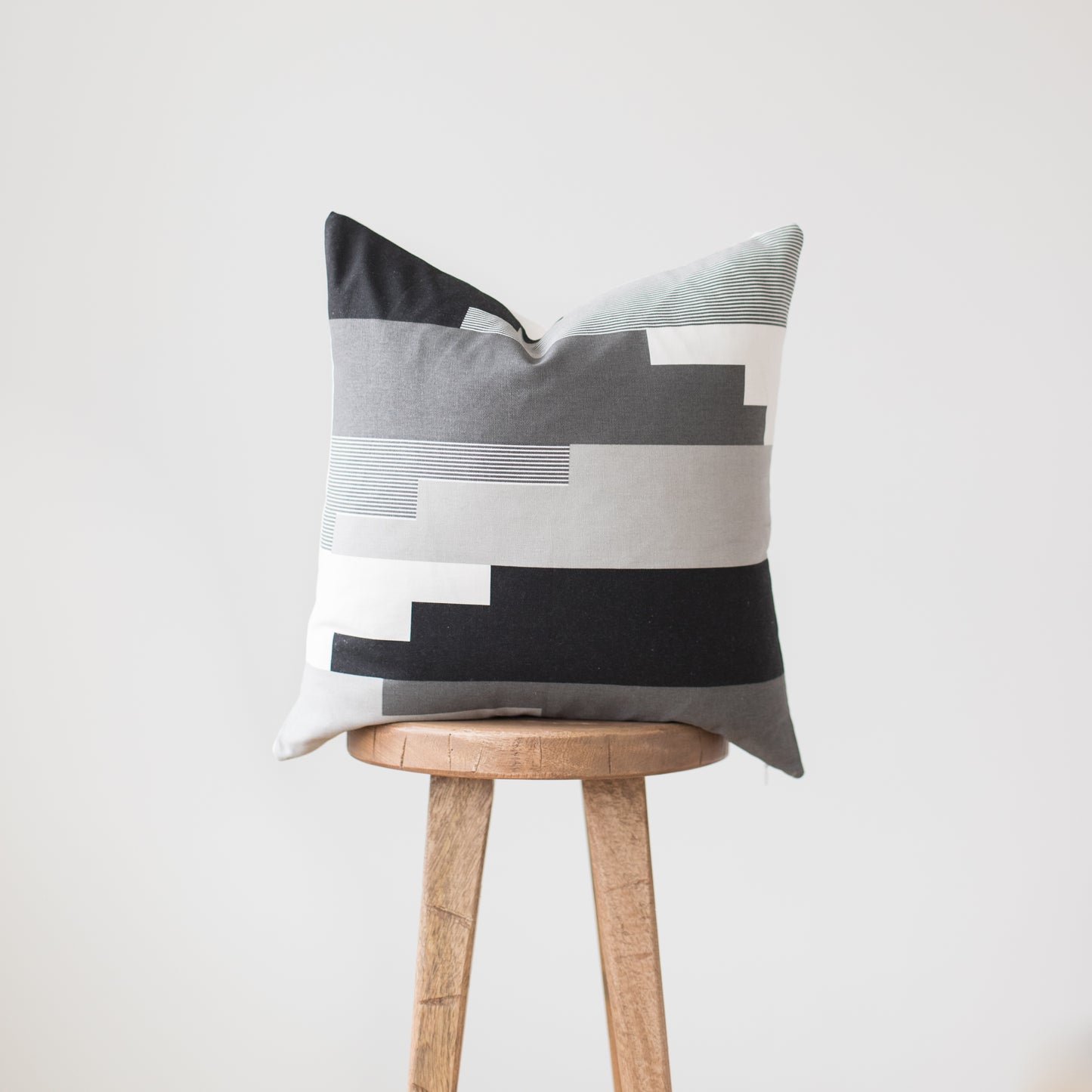 Throw pillow with geometric color-block design in black, gray, white, and blue stripes on wooden stool.