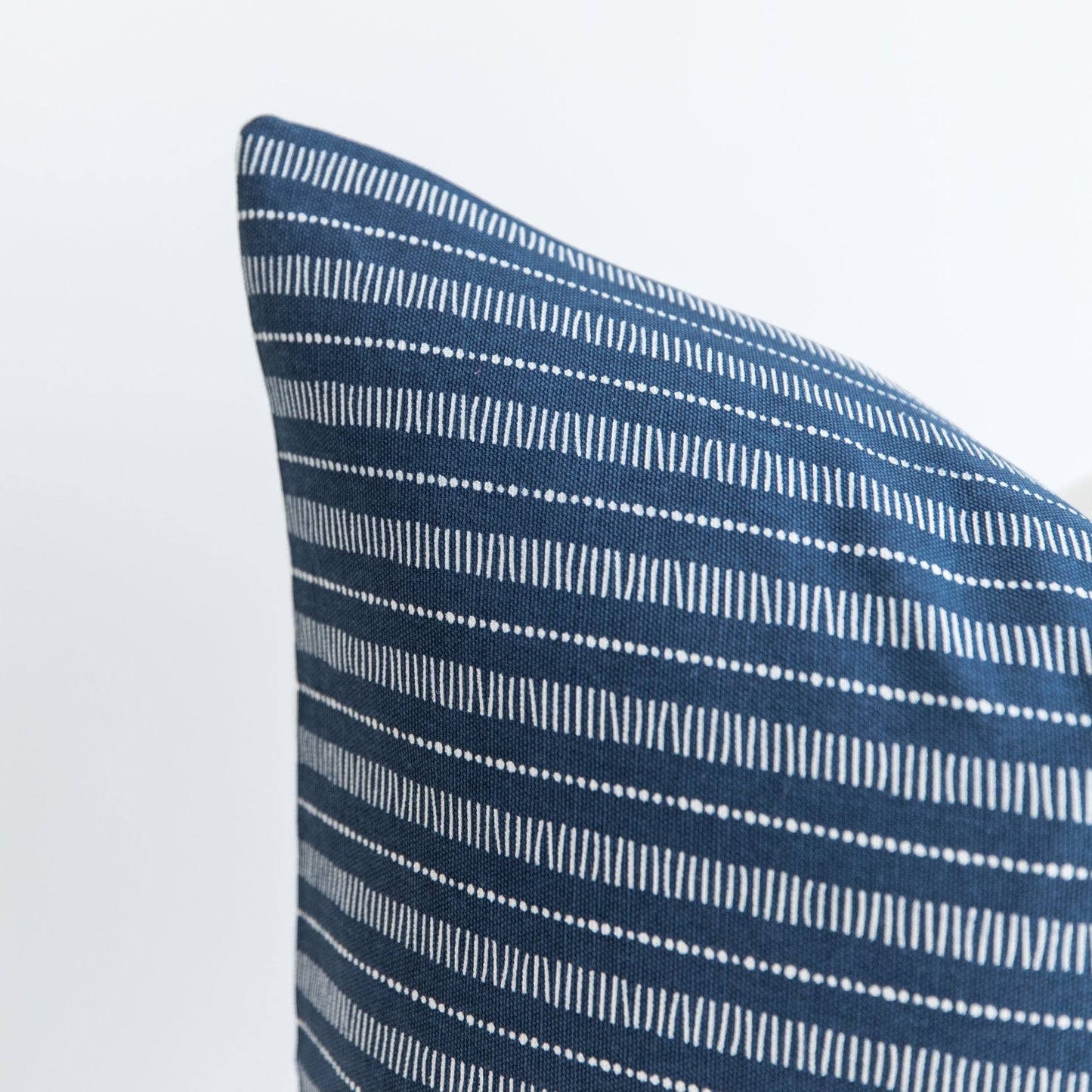 Blue pillow with white striped and dotted geometric pattern, shown at an angle against white background.