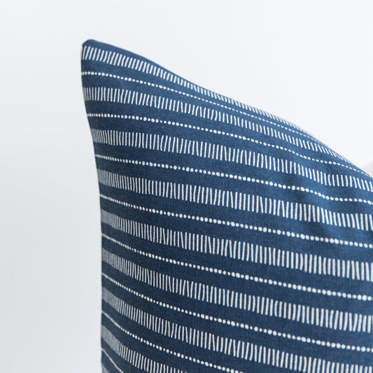 Blue pillow with white striped and dotted geometric pattern, shown at an angle against white background.