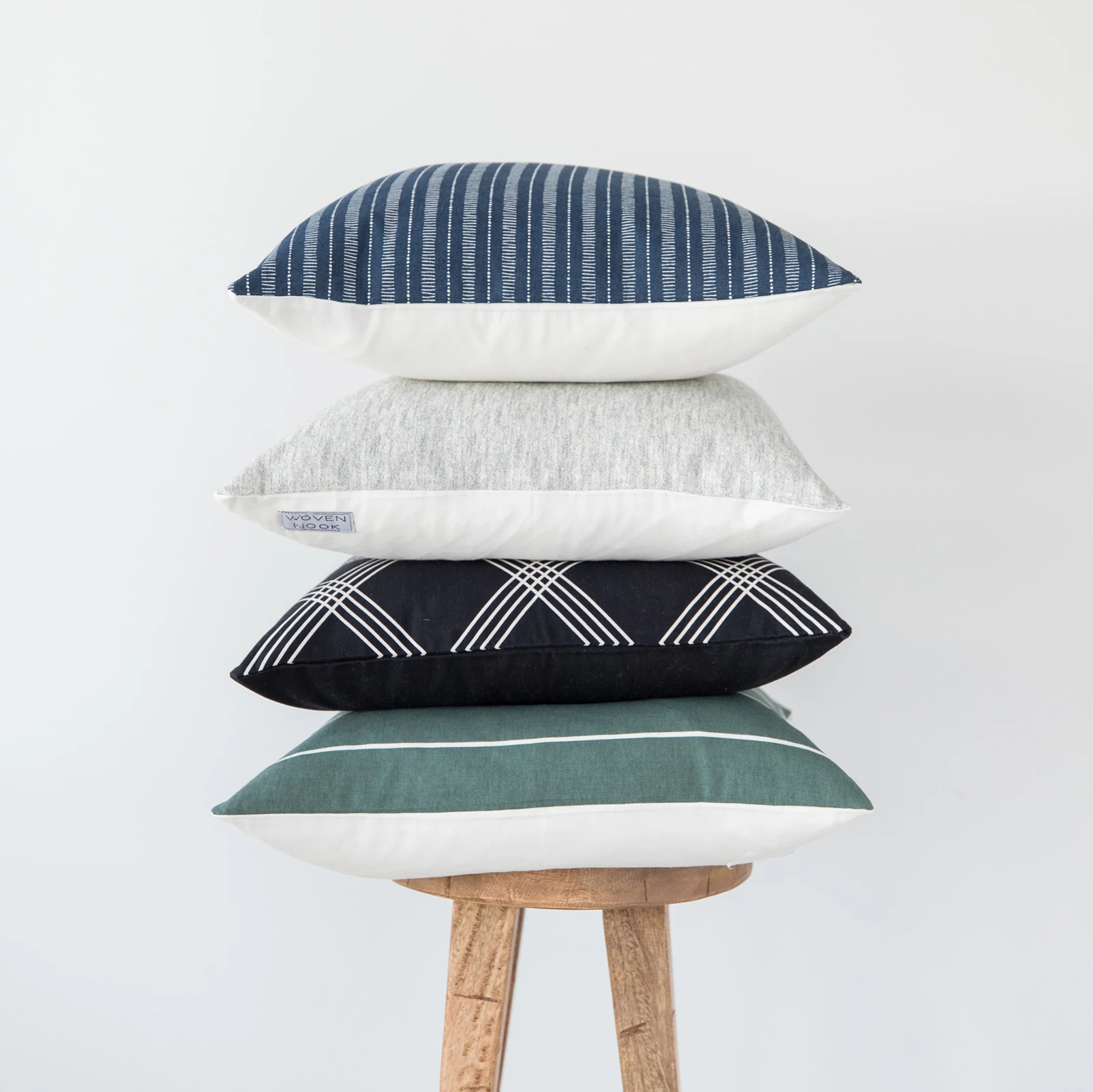 Four 18-inch pillows stacked on a wooden stool with blue striped, gray, black geometric, teal, and cream covers.
